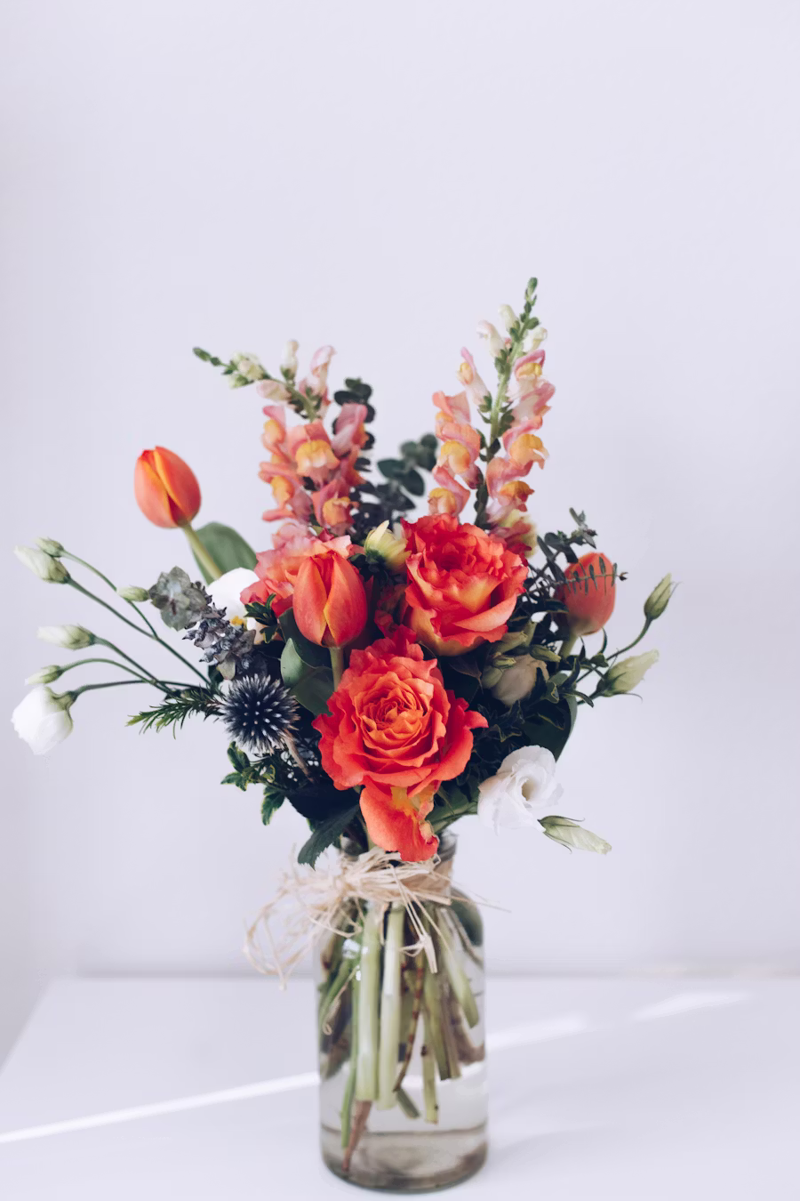 Composition libre dans un bocal en verre, roses orange vif, snapdragons saumon et chardons bleus